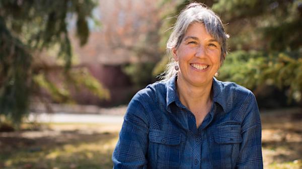 Ellen casually stands and smiles, light trees in the background.